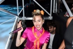 麦莉·赛勒斯（Miley Cyrus）梳高马尾，脖间系橘色丝巾，戴粉色亮钻猫眼墨镜，身穿粉色翻领连体裤