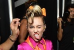 麦莉·赛勒斯（Miley Cyrus）梳高马尾，脖间系橘色丝巾，戴粉色亮钻猫眼墨镜，身穿粉色翻领连体裤